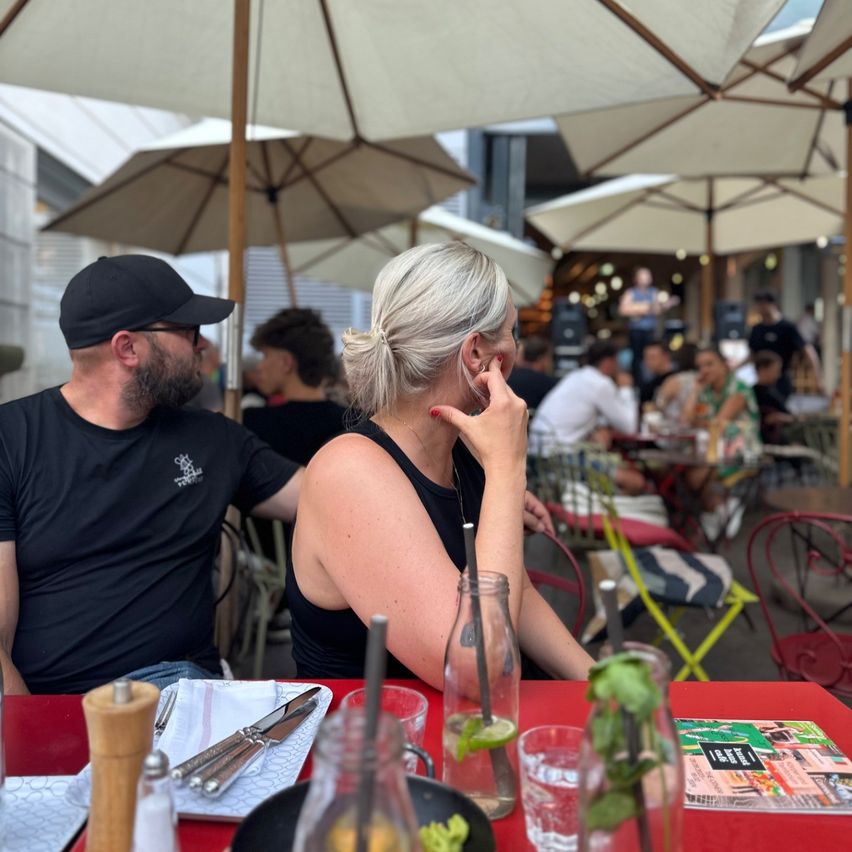 Ein Outdoor-Café mit weißen Sonnenschirmen, ein Mann und eine Frau sitzen an einem Tisch mit Getränken und Essen. Die Frau hat die Hand am Kinn und sieht nachdenklich aus.