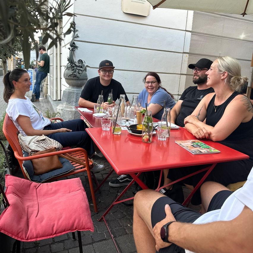 Eine Gruppe von Menschen sitzt um einen roten Tisch im Freien. Sie tragen Freizeitkleidung und haben Getränke. Eine Frau lächelt und ein Mann trägt eine Brille. Hinter ihnen steht ein Gebäude mit einer dekorativen Statue.