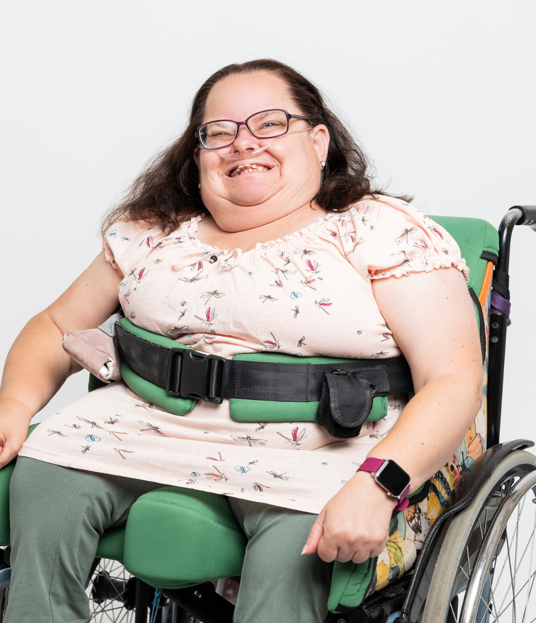 Eine Frau im Rollstuhl mit einem rosa Blumenblusen und grünen Hosen, lächelt mit einer Sauerstoffschlauch, trägt einen schwarzen Gürtel mit einer Tasche und eine rosa Uhr.
