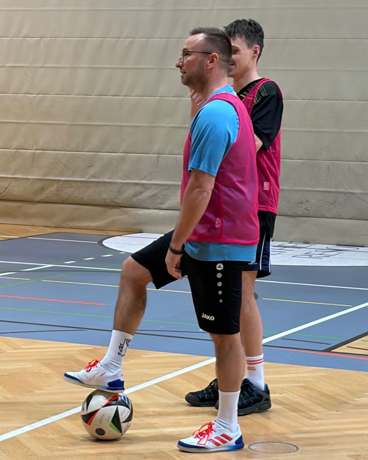 Zwei Männer in Sportkleidung stehen auf einem Spielfeld, einer tritt einen Fußball. Der Mann vorne trägt eine rosa Weste, der andere eine rote Weste. Sie tragen beide Turnschuhe.