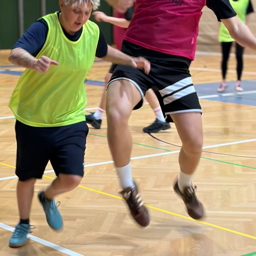 Zwei Spieler spielen auf einem Hallenspielfeld. Einer trägt eine neongrüne Weste und blaue Turnschuhe, der andere ein rotes und schwarzes Trikot mit braunen Schuhen.