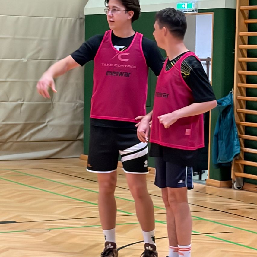 Zwei junge Männer in roten Westen stehen auf einem Basketballplatz und unterhalten sich. Der linke Mann gestikuliert.