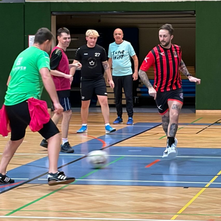 Fünf Männer spielen Hallenfußball. Ein Mann jagt einem Ball hinterher, ein anderer steht hinter ihm. Ein Mann in einem blauen Shirt und zwei andere schauen zu.