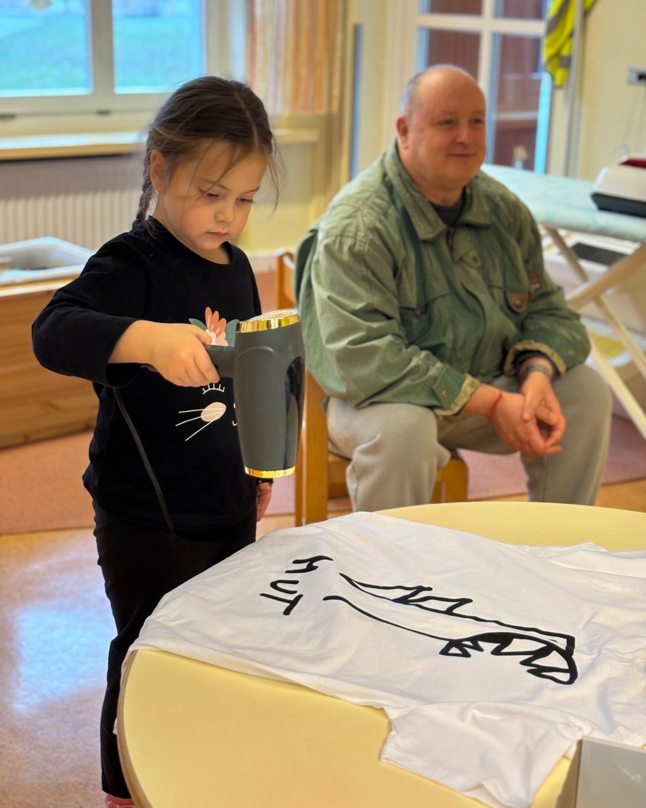 Ein junges Mädchen benutzt einen Haartrockner auf einem T-Shirt in einem Raum. Ein älterer Mann sitzt in der Nähe und lächelt.