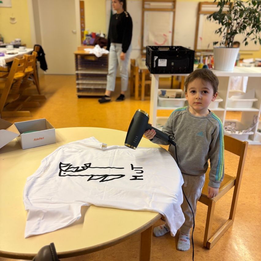 Ein kleiner Junge benutzt einen Föhn, um ein weißes T-Shirt auf einem Tisch zu trocknen, während eine Frau im Hintergrund vorbeigeht.