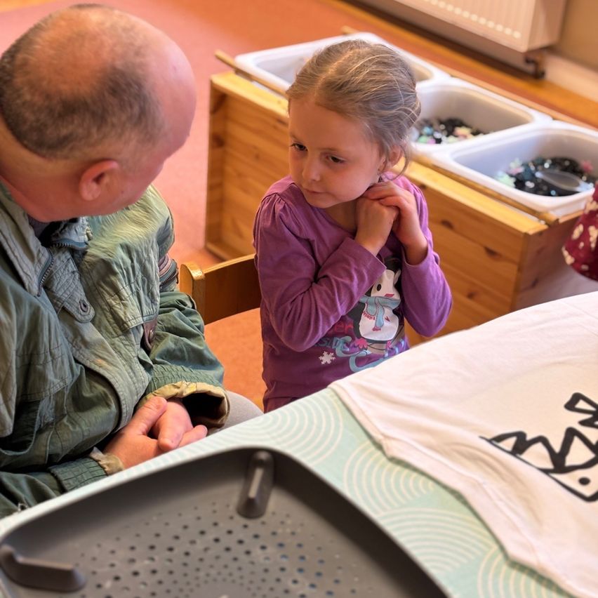 Ein Mann und ein junges Mädchen sitzen zusammen. Der Mann trägt eine grüne Jacke. Das Mädchen trägt ein lila Langarmshirt mit Schneemann-Design. Es gibt einen Tisch mit einem weißen Shirt darauf.