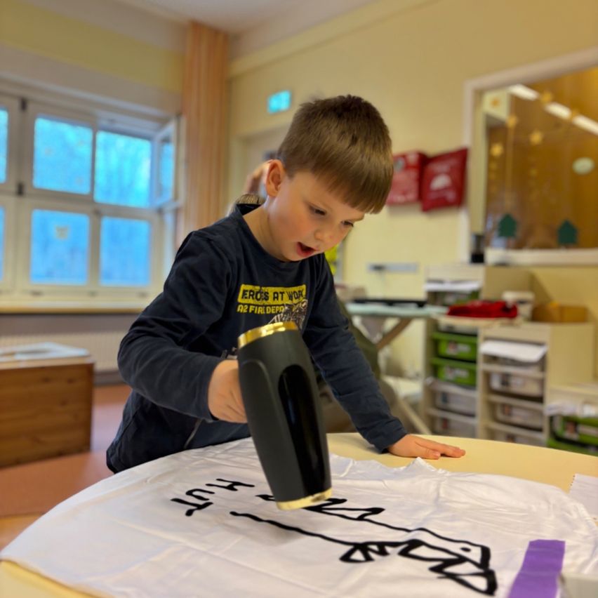 Ein junger Junge benutzt ein schwarzes Bügeleisen auf einem weißen Tuch mit einer Dinosaurier-Zeichnung darauf. Er befindet sich in einem Klassenzimmer mit gelben Wänden und großen Fenstern.