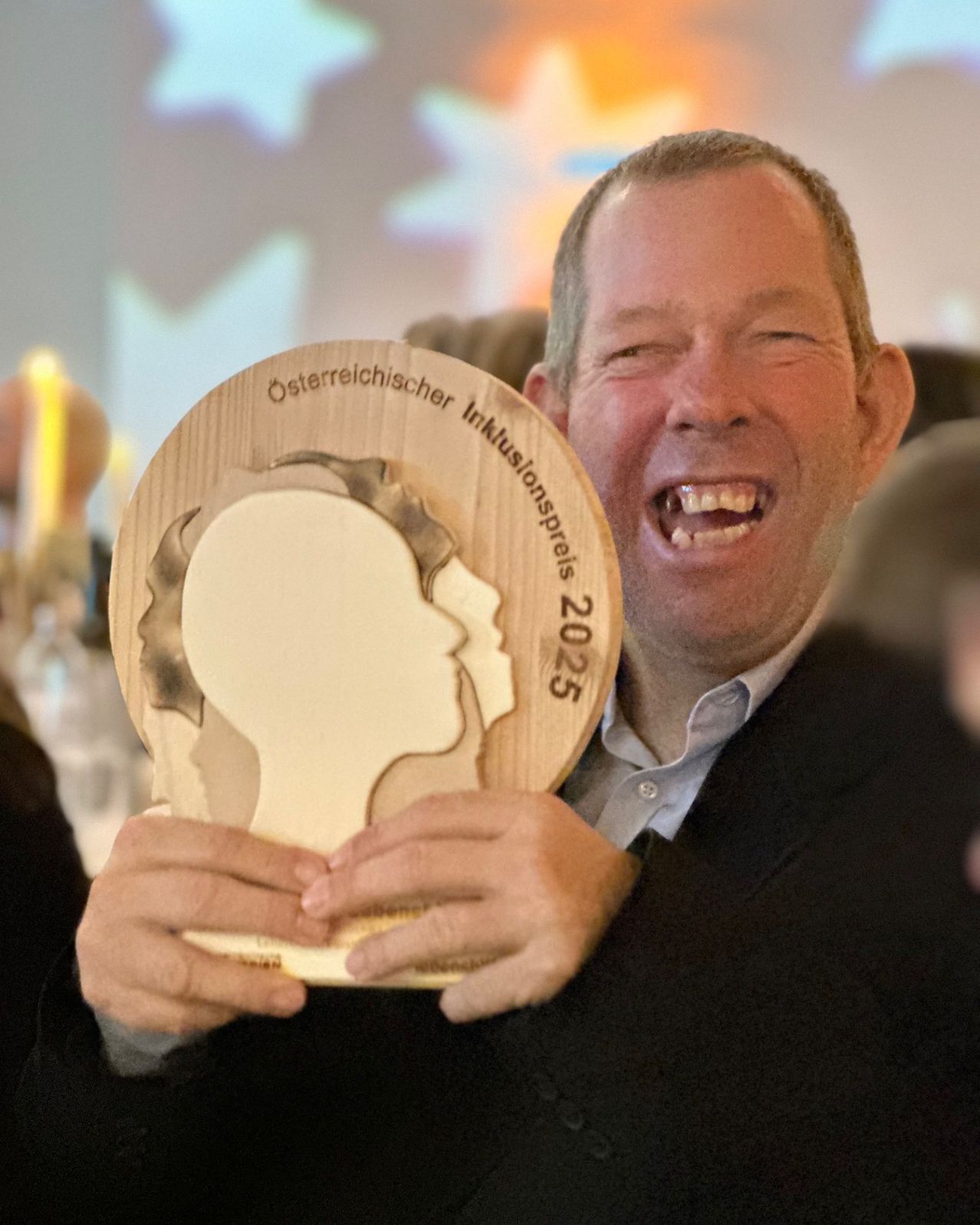Ein Mann hält eine hölzerne Plakette mit der Aufschrift 'Osterreichischer Innovationspreis 2025'. Er lächelt mit offenem Mund, vielleicht bei einer Zeremonie.