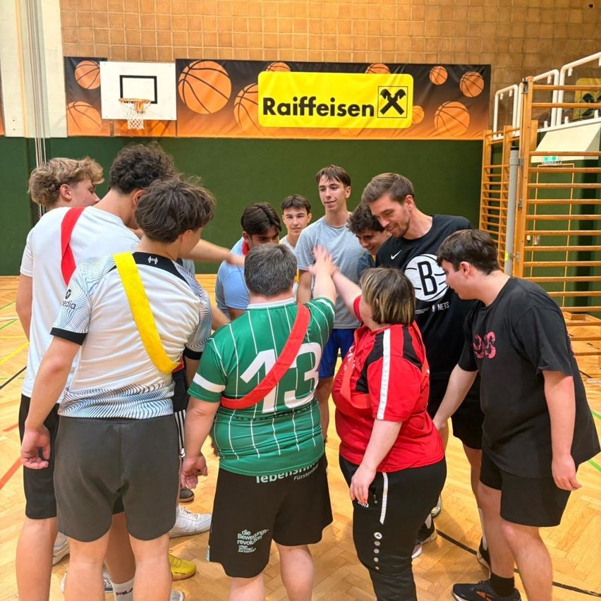 Eine Gruppe junger Menschen und Erwachsene versammelt sich in einer Sporthalle, wahrscheinlich für ein Basketballtraining. Eine Person in einem grünen Trikot mit der Nummer 13 an der Spitze leitet die Gruppe.