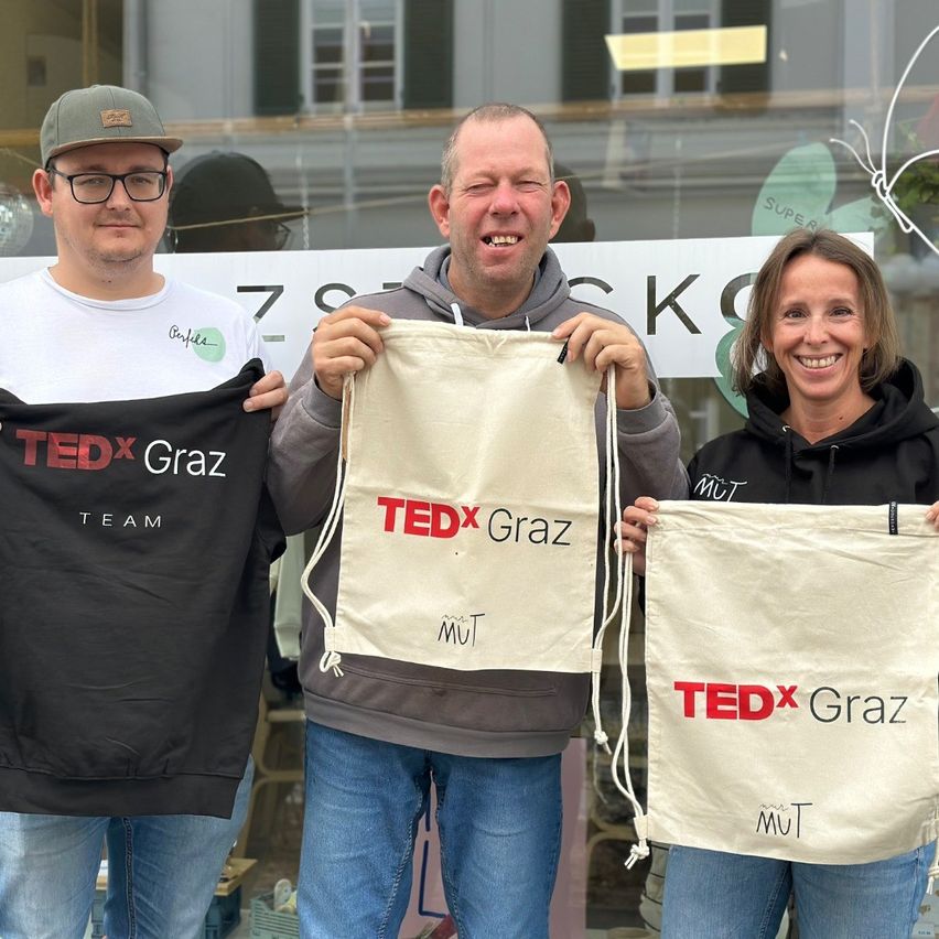 Drei Menschen stehen eng beieinander und lächeln. Der Mann auf der linken Seite trägt ein schwarzes T-Shirt mit 'TEDx Graz Team'. Die Person in der Mitte hält eine weiße Tasche mit 'TEDx Graz'. Die Frau auf der rechten Seite hält eine beige Tasche mit 'TEDx Graz'. Sie stehen vor einer Glasscheibe mit 'K'.