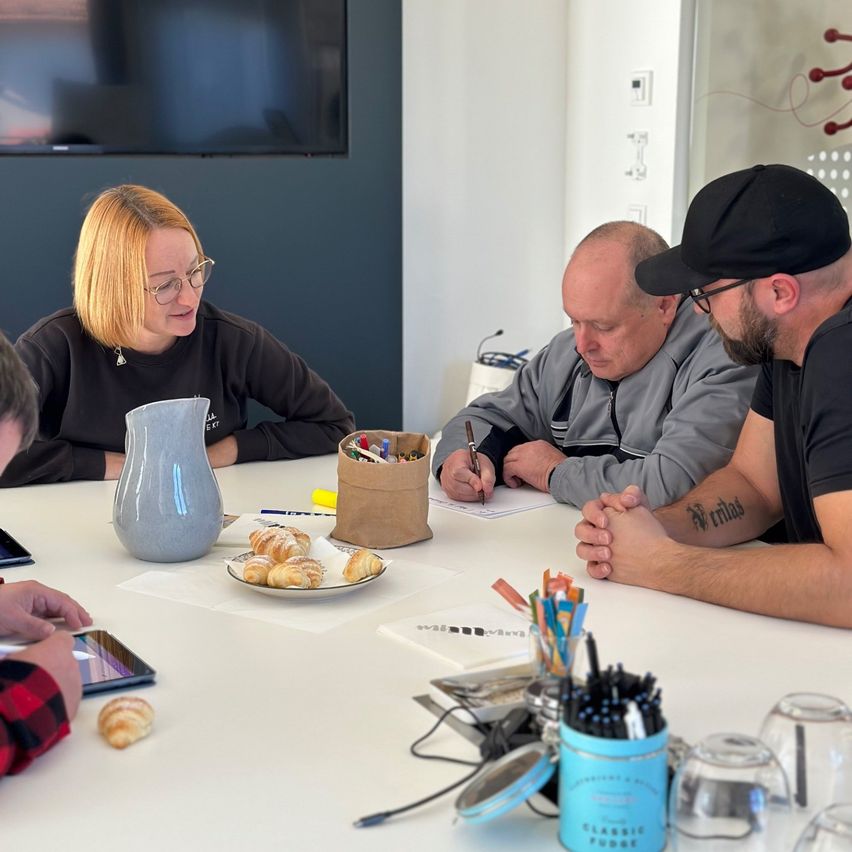 Eine Gruppe von Menschen sitzt um einen Tisch herum und ist in ein Meeting vertieft. Ein Mann schreibt auf Papier, während ein anderer Mann mit einem Tattoo auf seinem Arm ihn ansieht. Eine Frau mit Brille und einer Halskette sitzt in der Nähe. Es gibt Croissants, Stifte und ein Tablet auf dem Tisch.