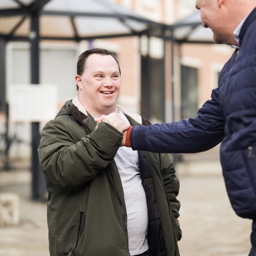 Ein Mann in einer grünen Jacke lächelt, während er einem anderen Mann in einer blauen Jacke die Hand schüttelt. Sie stehen vor einem Gebäude.