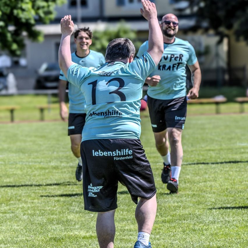 Drei Personen in identischen blauen Trikots laufen auf einem Rasenfeld. Die Person in der Mitte, die die Nummer 13 trägt, hebt beide Hände.