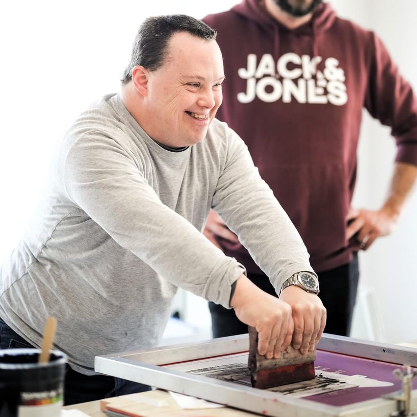 Ein Mann lächelt, während er an einem Siebdruckprojekt arbeitet. Ein anderer Mann in einer Jack & Jones Kapuzenjacke in Weinrot steht hinter ihm.