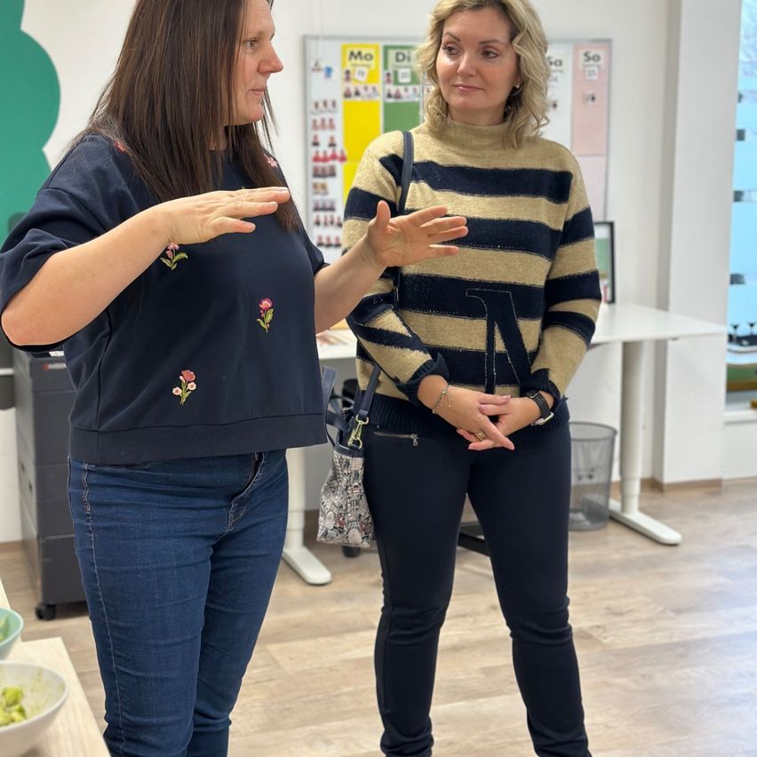 Two women stand in an office, engaged in conversation. The woman on the left wears a navy blue shirt with floral embroidery. The woman on the right wears a striped sweater. Behind them, there are white desks, a bulletin board, and a trash bin.