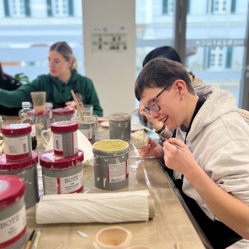 Several people sit around a table covered with paint cans, cups, and paper towels. A woman smiles while holding a paintbrush.