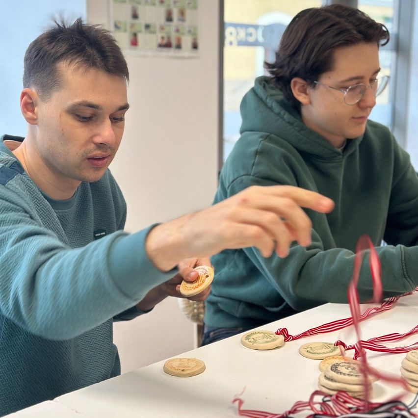 Zwei Männer sitzen an einem Tisch, der mit Keksen und Bändern bedeckt ist. Ein Mann zeigt mit der Hand, während der andere aufmerksam zusieht. Sie nehmen wahrscheinlich an einer Keksdekorationsaktivität teil.
