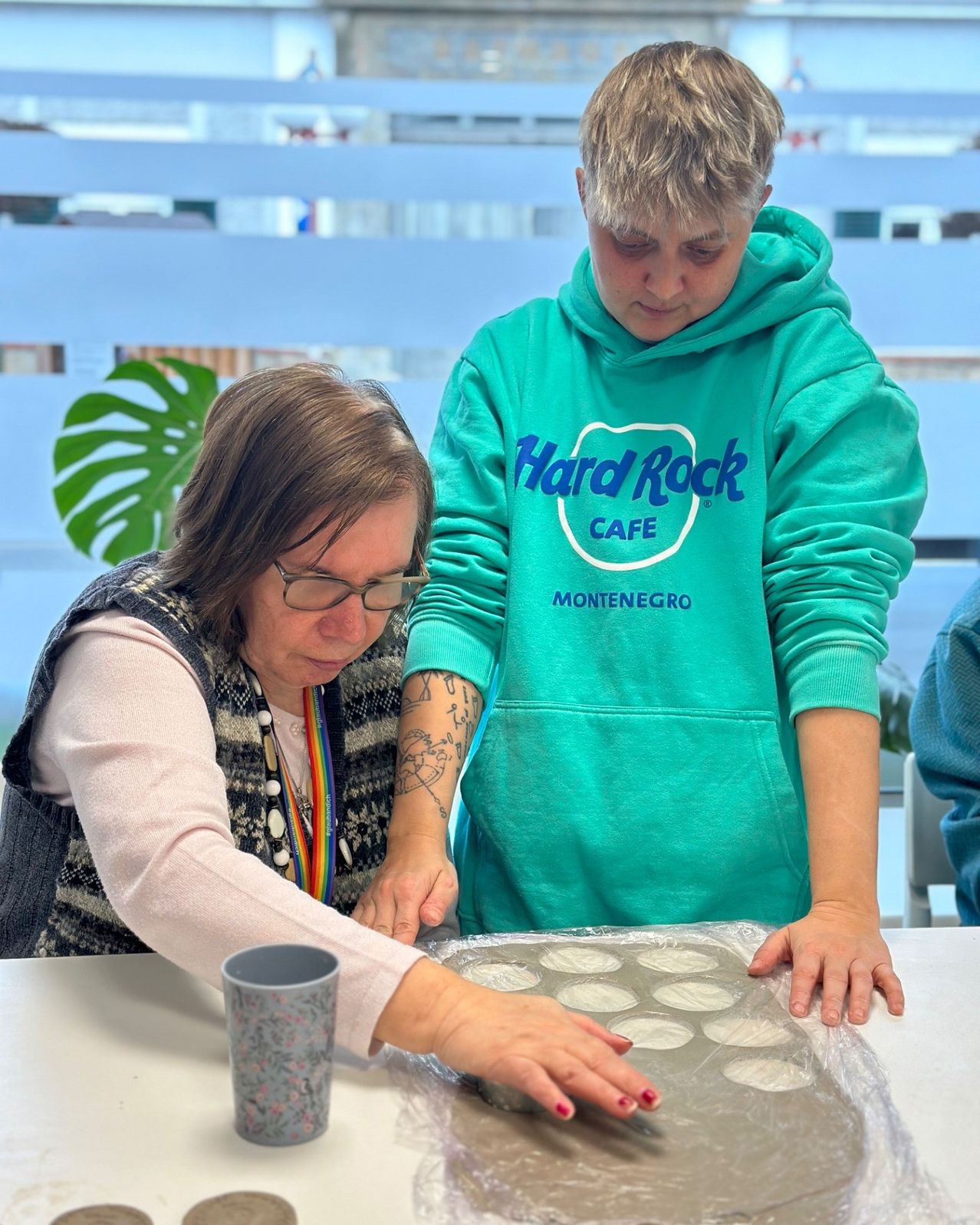 Zwei Frauen stehen über einem Tisch mit einem Muffinblech, eine trägt eine Hard Rock Cafe-Hoodie und die andere trägt eine Weste mit einem Regenbogenhalsband.