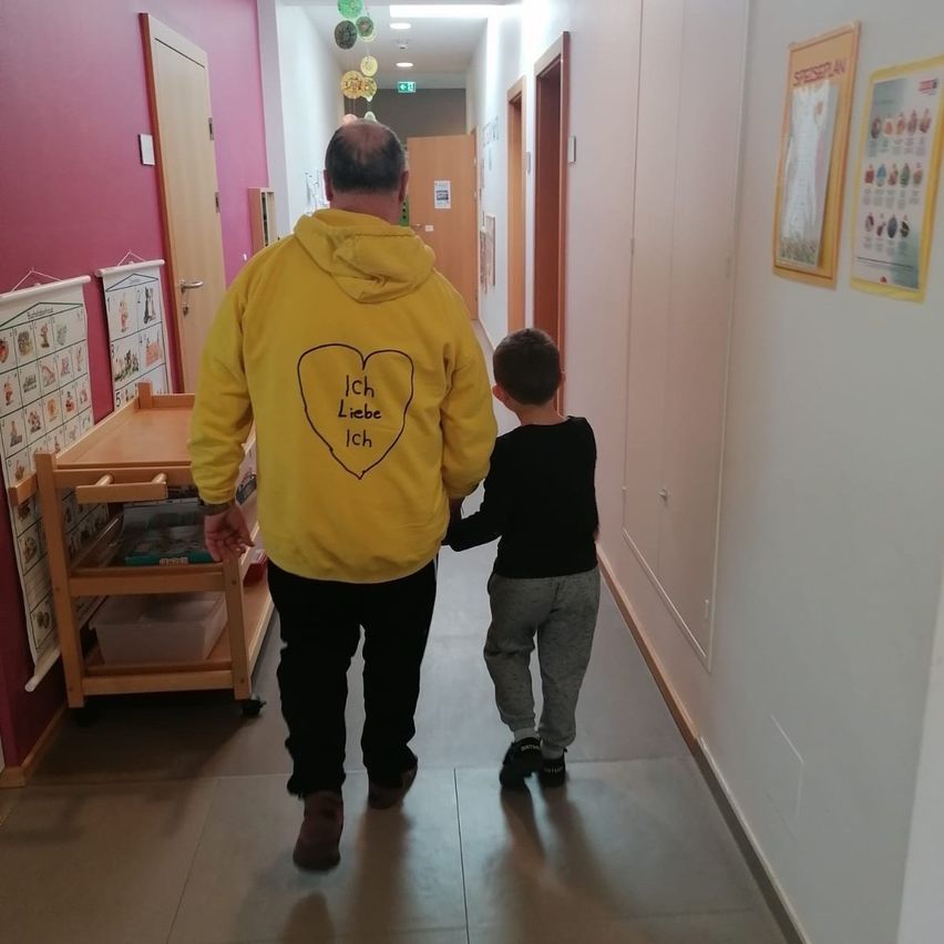 An adult male wearing a yellow hoodie walks with a young boy down a hallway. They are holding hands.