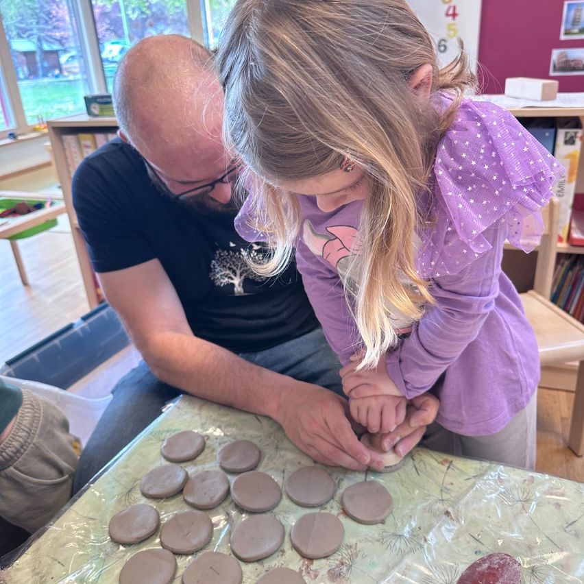 A man and a girl are crafting clay circles on a table, with a child nearby. A bookshelf is behind them, and windows show a grassy yard.