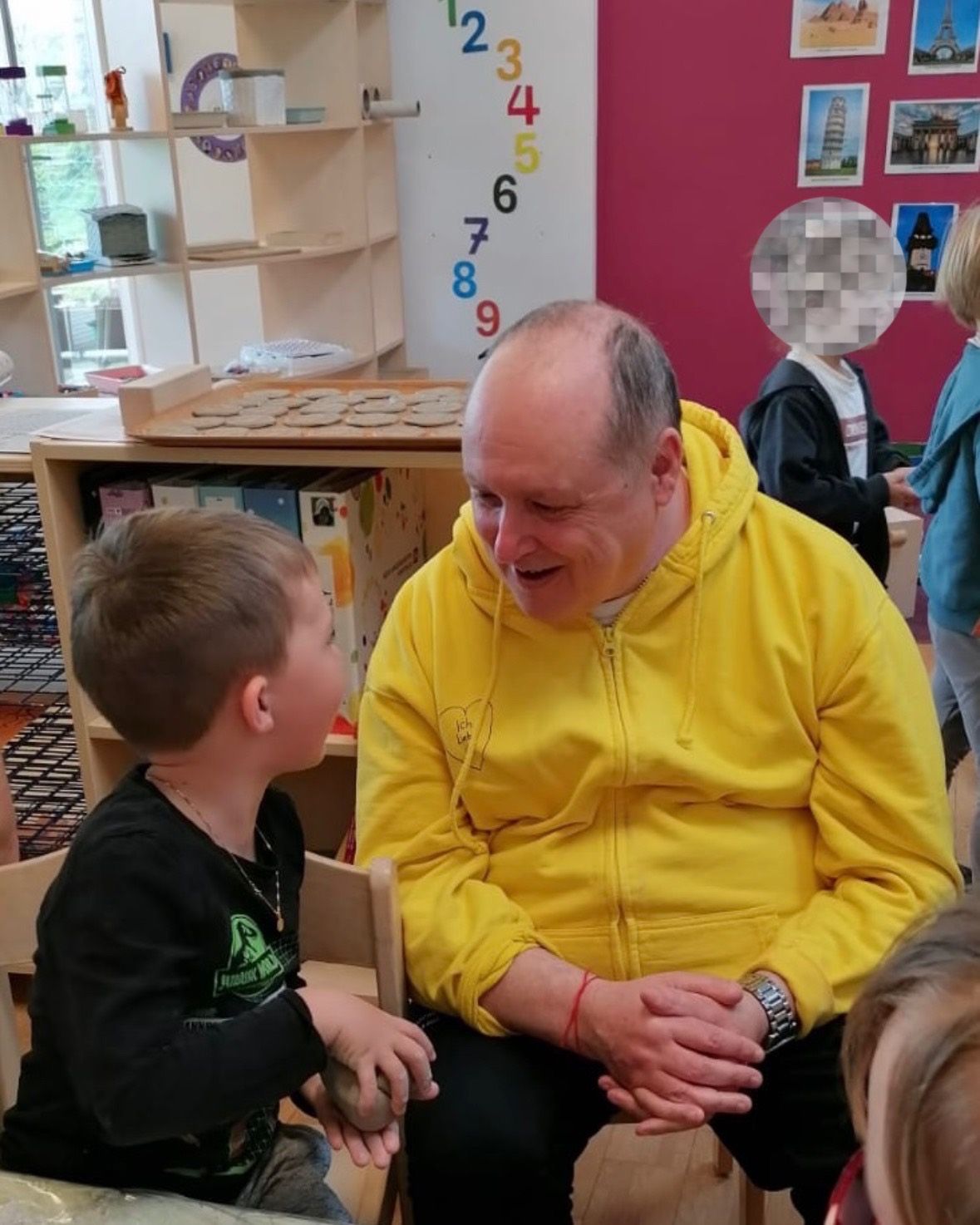 A man in a yellow hoodie is sitting in a classroom and smiling at a young boy in a black shirt. Behind them is a blurred figure. The classroom has shelves with books and other items.