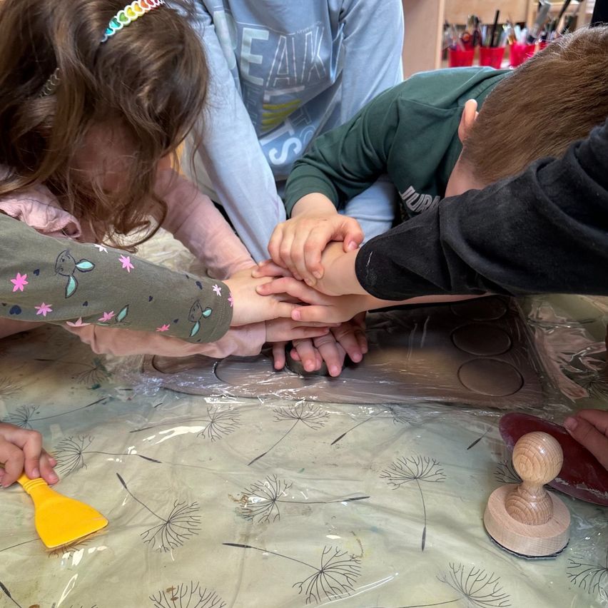 Several children are gathered around a table covered in plastic. They are holding hands and appear to be doing an art project. One child is stamping a wooden stamp.