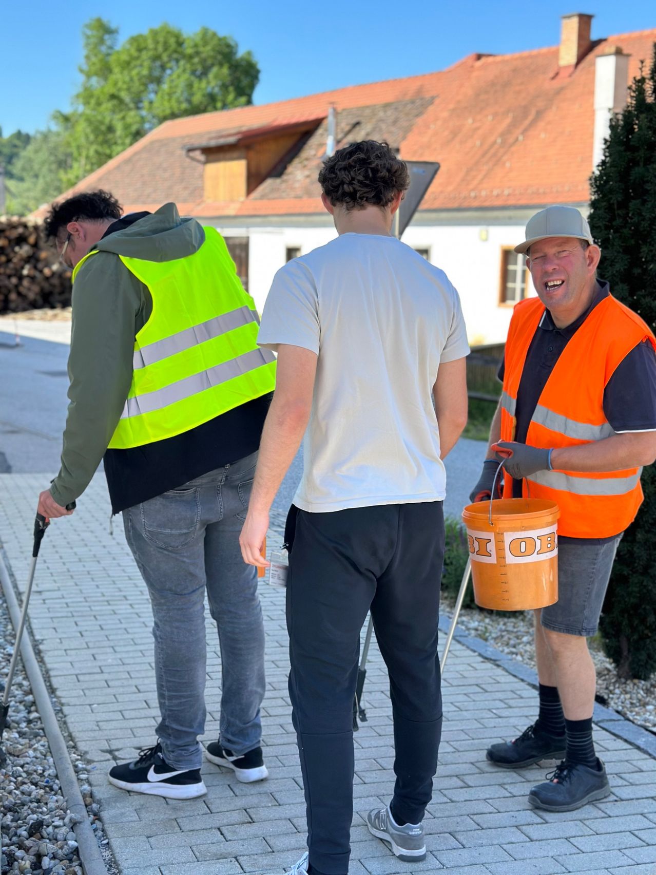 Three people are outside. One holds a bucket. Two wear high visibility vests. One wears a hat.