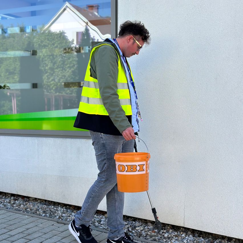 A man wearing a safety jacket is carrying an orange bucket labeled OBI on the sidewalk. He seems to be looking at something on the wall.