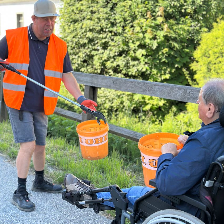 A man in an orange vest holds a tool and gestures while standing next to a man in a wheelchair who holds a bucket. They are on a road with a fence and trees in the background.