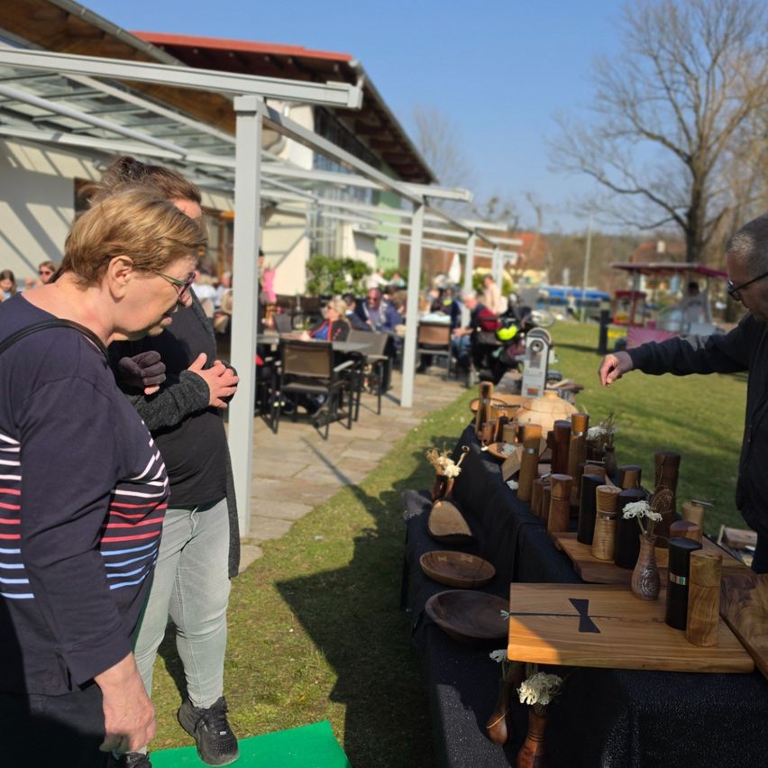 Menschen versammeln sich um einen Tisch mit Holzgegenständen unter einem Vordach an einem sonnigen Tag. In der Nähe sitzen Personen an Tischen und Stühlen, und eine andere Person zeigt auf den Tisch. Ein Gebäude mit rotem Dach ist im Hintergrund.