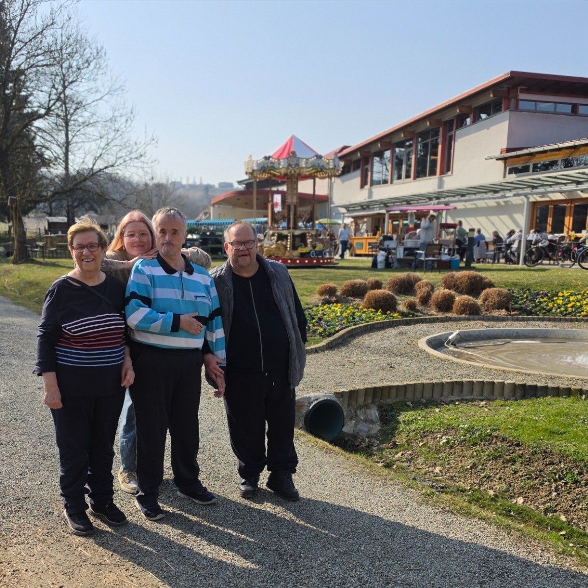 Vier Personen posieren für ein Foto in einem Park. Sie stehen auf einem Kiesweg. Dahinter befindet sich ein Gebäude mit einer Karussellfahrt. Bäume und Pflanzen sind in der Umgebung.