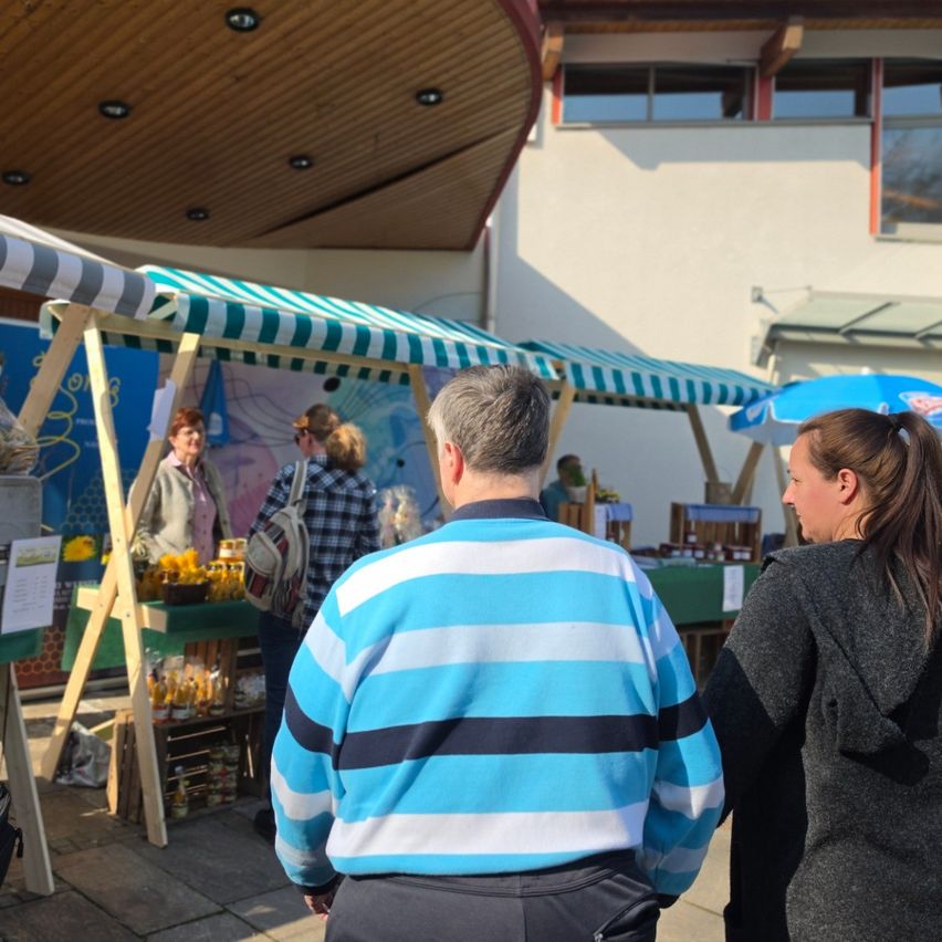 Auf einem Markt im Freien unterhält sich ein Mann in einem blau-weiß gestreiften Pullover mit einer Frau. Hinter ihnen zeigen Stände verschiedene Waren an, einer mit gelben Blumen. Die Szene spielt sich unter einem gewölbten Dach ab, in der Nähe eines Gebäudes mit Glasfenstern.