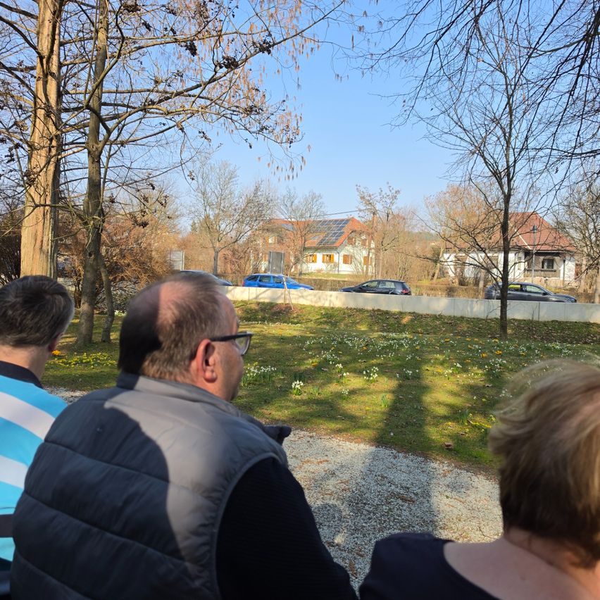 Drei Personen sitzen in einem Park und blicken auf eine Rasenfläche mit Häusern und geparkten Autos. Der Himmel ist klar und blau.