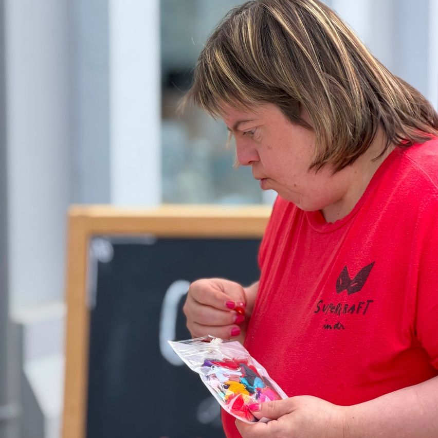 Eine Frau mit kurzen blonden Haaren, die ein rotes T-Shirt trägt, schaut auf einen Plastikbeutel voller bunter Perlen. Sie steht vor einer hölzernen Tafel.