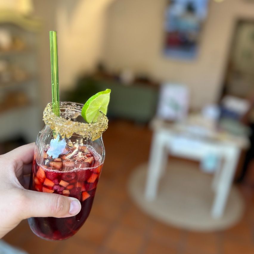 A hand holding a glass filled with a red drink, garnished with a lime slice and a green straw. The background shows a blurry room with a white table and a rug.