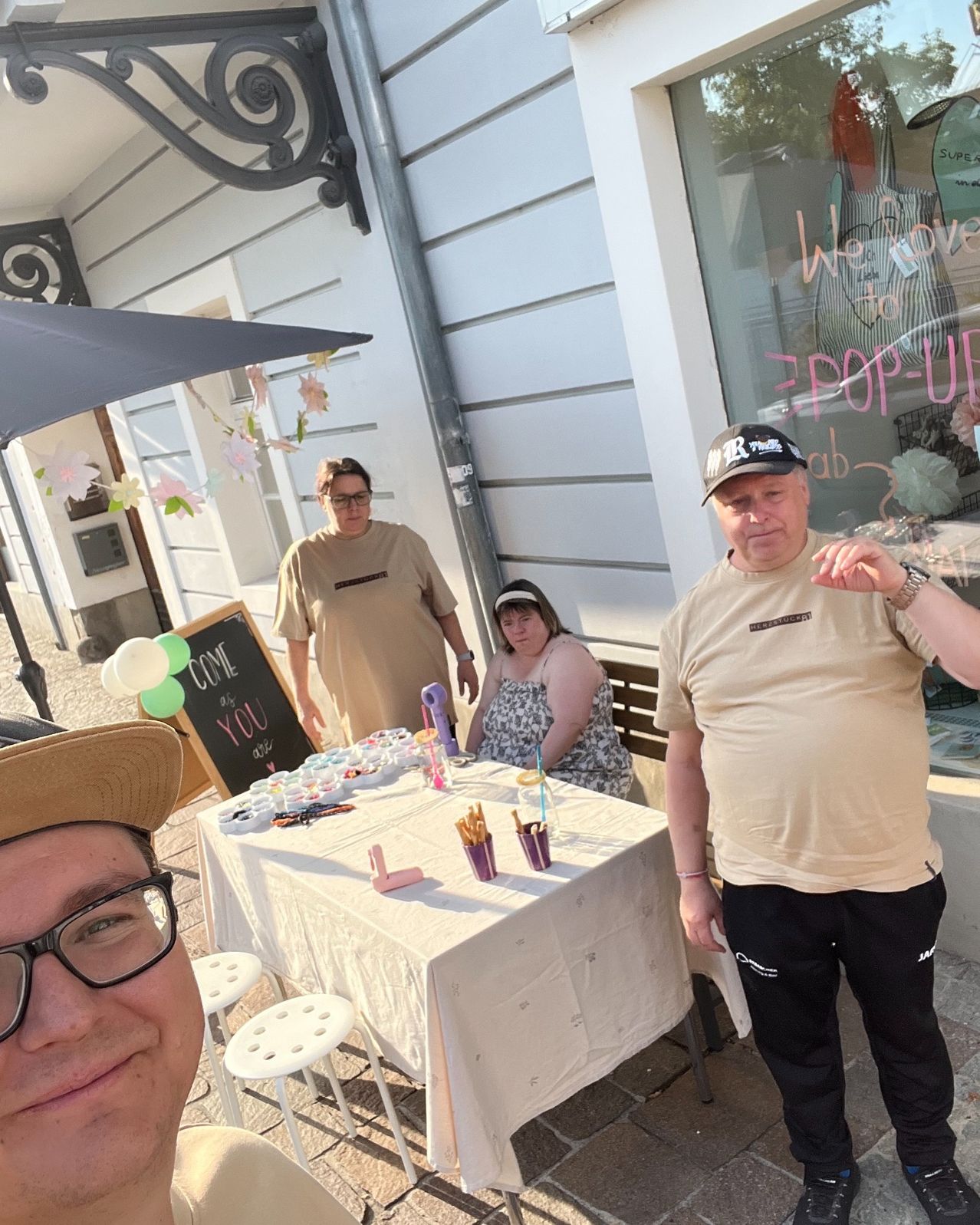 A group of people stands outside a shop, with a table full of crafts and decorations. A woman sits at the table, while a man stands nearby. A blackboard with a message is visible.