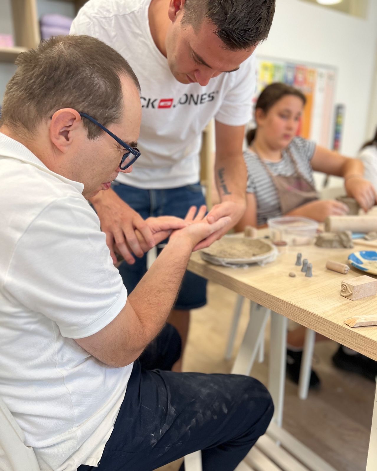 Ein Mann mit Brille und weißem T-Shirt arbeitet an einem Tisch an einem Tonprojekt, während eine Frau in der Nähe ist.