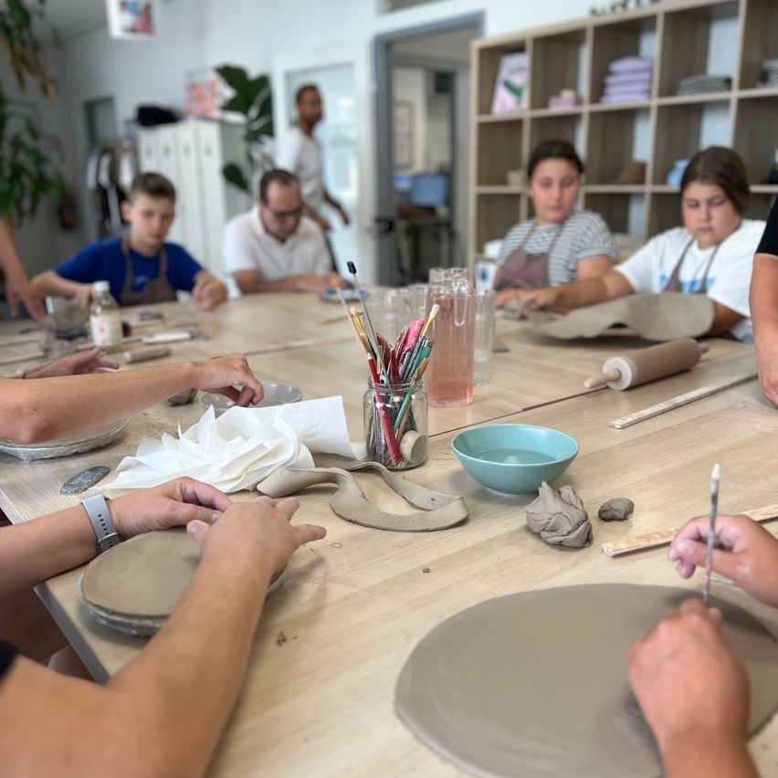 Eine Gruppe von Menschen sitzt um einen Tisch herum und ist in einer Töpferaktivität engagiert. Sie verwenden Ton und verschiedene Werkzeuge.