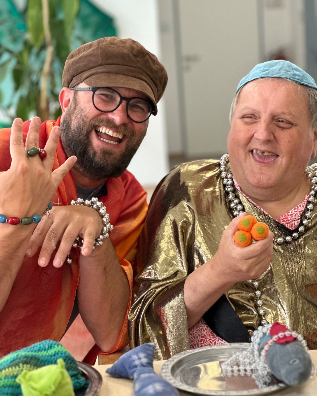 Ein Mann und eine Frau lächeln und posieren für ein Foto. Der Mann trägt einen Hut und eine Brille. Die Frau trägt einen goldenen Umhang und hält orangefarbene Süßigkeiten.