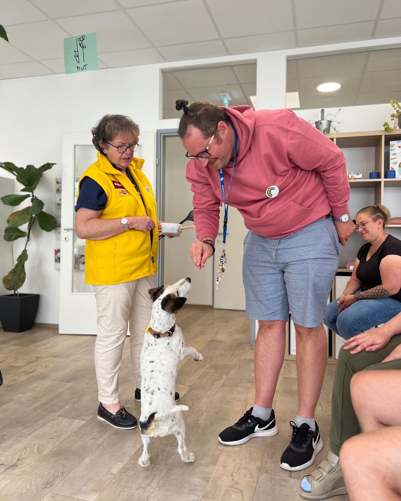 Eine erwachsene Frau in einer gelben Weste steht neben einem Mann in einem rosa Hoodie, beide schauen einen Hund an, der auf seinen Hinterbeinen steht. Eine Frau sitzt im Hintergrund auf einem Sofa.