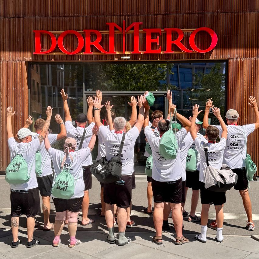 Eine Gruppe von Menschen steht vor einem Gebäude mit dem Namen 'DORTMERO'. Sie tragen weiße T-Shirts mit grünen Taschen und manche heben ihre Hände.