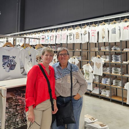 Zwei Frauen stehen vor einem Laden voller T-Shirts. Sie lächeln und scheinen für ein Foto zu posieren. Hinter ihnen hängen Regale mit T-Shirts.