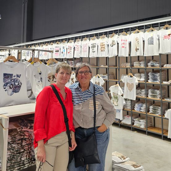 Zwei Frauen stehen vor einem Laden voller T-Shirts. Sie lächeln und scheinen für ein Foto zu posieren. Hinter ihnen hängen Regale mit T-Shirts.