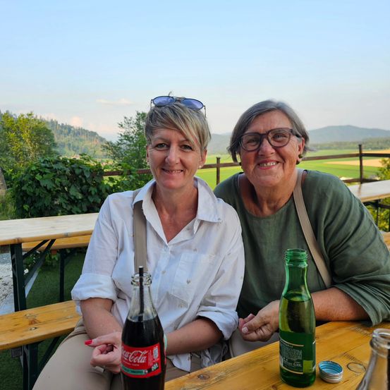 Zwei Frauen sitzen lächelnd auf einer Bank im Freien. Eine hält eine Coca-Cola-Flasche, die andere eine grüne Flasche. Sie sind von Holztischen umgeben und vor einer landschaftlichen Kulisse.