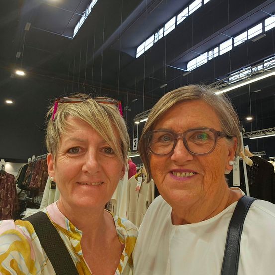 Zwei Frauen stehen nebeneinander und lächeln für ein Foto in einem Bekleidungsgeschäft. Eine hat eine Brille auf dem Kopf und die andere trägt eine Brille. Sie stehen vor einer Ausstellung von Kleidungsstücken.