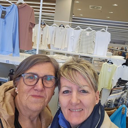 Zwei Frauen lächeln in einem Bekleidungsgeschäft. Verschiedene Hemden hängen an Kleiderständern hinter ihnen. Die linke Frau trägt eine Brille und eine braune Jacke, während die rechte Frau einen blauen Schal hat.