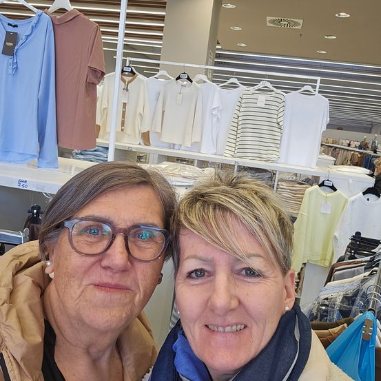 Zwei Frauen lächeln in einem Bekleidungsgeschäft. Verschiedene Hemden hängen an Kleiderständern hinter ihnen. Die linke Frau trägt eine Brille und eine braune Jacke, während die rechte Frau einen blauen Schal hat.