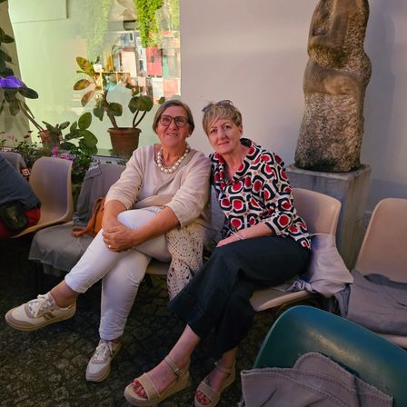 Zwei Frauen sitzen auf Stühlen in einem Raum und lächeln für ein Foto. Hinter ihnen steht eine große Steinstatue und ein Fenster mit Pflanzen.