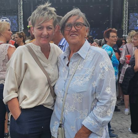 Zwei Frauen stehen lächelnd für ein Foto bei einem Outdoor-Event mit einer großen Bühne im Hintergrund. Leute gehen herum und einige schauen zur Bühne.