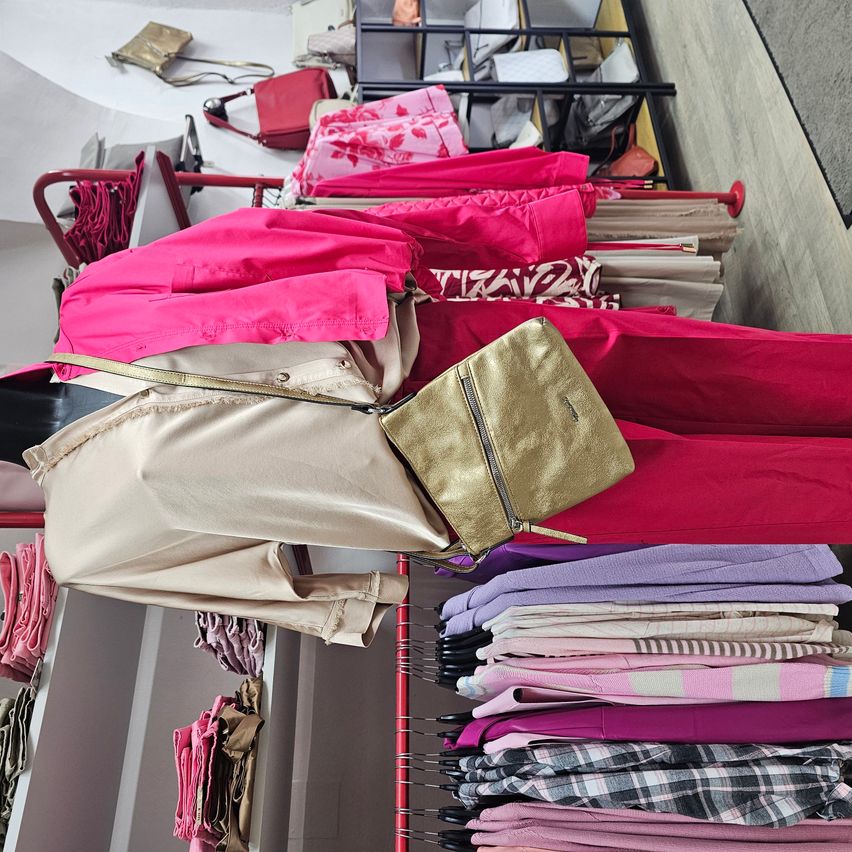 A clothing store displays a variety of items, including pink and beige pants, a pink jacket, and a gold handbag. Shelves and racks are filled with folded clothes and handbags, showcasing a range of colors and patterns.